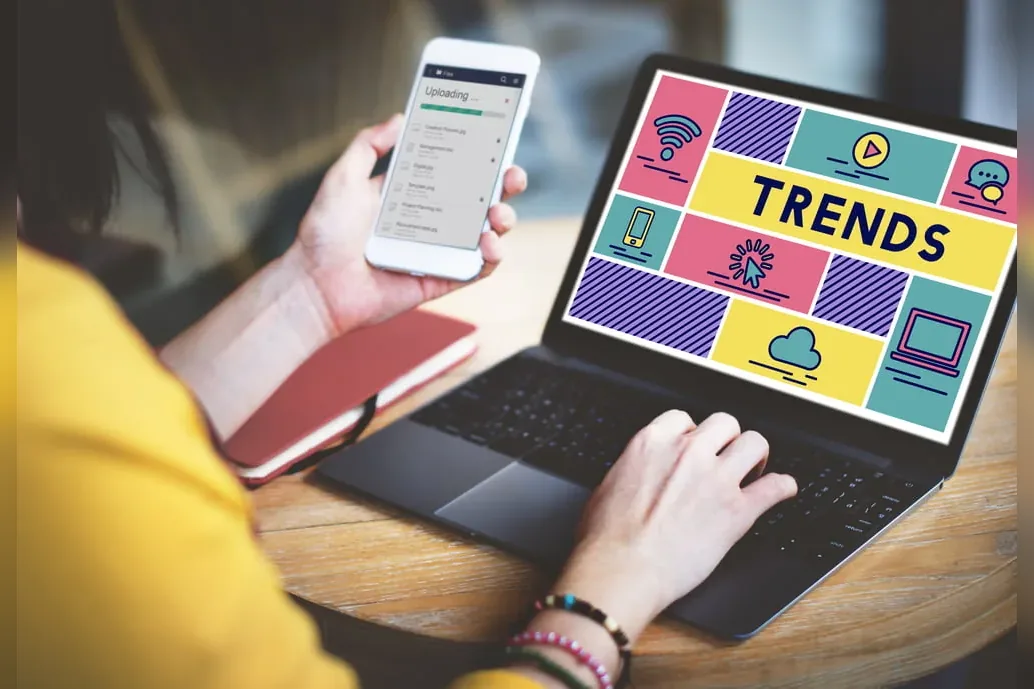 Someone at a laptop showing a grid of technology icons headed Trends, holding a phone in the other hand.
